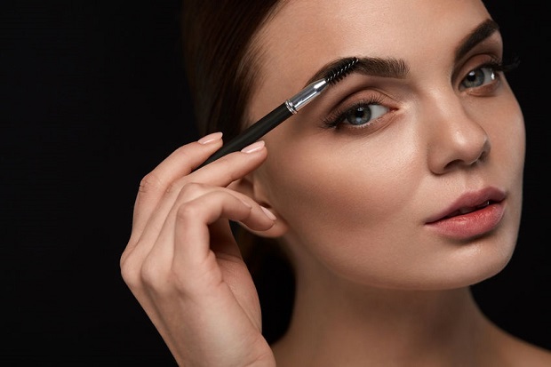 Eyebrows Brushing. Closeup Of Beautiful Young Female Model Face With Professional Makeup And Brow Brush On Black Background. Sexy Woman Brushes Perfect Brows. Beauty Cosmetics. High Resolution Image
